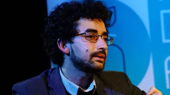 Homme avec des lunettes, cheveux bouclés, en discussion, vêtu d'une veste.