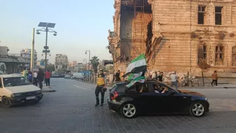 Una strada con una costruzione danneggiata, persone e un'auto con una bandiera.