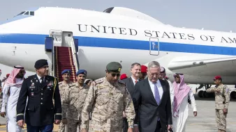 The image depicts a group of individuals exiting from a large aircraft marked with the words "UNITED STATES OF AMERICA." In the foreground, there are several military personnel and officials. One individual appears to be dressed in a military uniform, while others are in formal attire. They seem to be engaged in conversation as they walk down the steps from the plane. The backdrop includes the aircraft and an airport setting, suggesting a diplomatic visit or military engagement.