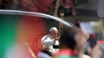 L'image montre une scène d'un événement public avec le Pape François. Il est souriant et fait un geste de la main en saluant la foule. On peut apercevoir des drapeaux multicolores dans le premier plan, ce qui indique une ambiance festive et dynamique. En arrière-plan, on distingue des illustrations ou des représentations qui semblent évoquer une célébration ou un rassemblement spirituel. L'ensemble dégage une atmosphère de joie et d'enthousiasme de la part des personnes présentes.