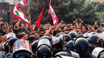 در این تصویر، یک تجمع اعتراضی در لبنان دیده می‌شود. گروهی از مردم در حال شعار دادن و به‌سوی دوربین نگاه می‌کنند، در حالی که پرچم‌های لبنان را در دست دارند. در مقابل آن‌ها، گروهی از نیروهای پلیس با لباس‌های ضدشورشی و کلاه‌خودهایی مشغول محافظت از جایگاه هستند. فضای تصویر تنش‌آلود و احساسات قوی در بین معترضین و نیروهای پلیس به وضوح حس می‌شود. در پس‌زمینه، ساختمان‌ها و درختان وجود دارند که به جو عمومی صحنه اشاره می‌کنند.