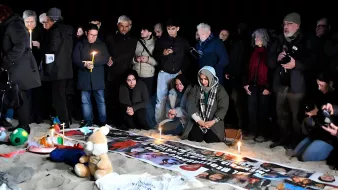 Un gruppo di persone in cerchio, con candele accese e striscioni, su una spiaggia al buio.