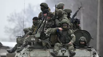 L'image montre un groupe de soldats armés, équipés de vêtements militaires et de casques, se tenant sur un véhicule blindé. Ils semblent en position attentive, probablement lors d'une opération militaire. L'ambiance est sérieuse et le paysage environnant est sombre, ce qui renforce le contexte militaire et opérationnel. Les soldats portent des masques et des lunettes, ce qui indique qu'ils sont préparés pour des conditions difficiles.