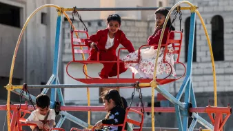 L'image montre des enfants jouant sur une grande structure de jeux colorée. On voit des balançoires fixées à une plateforme, dont certains enfants sont assis. Les enfants affichent des expressions joyeuses, profitant de leur temps de jeu. La scène se déroule dans un espace ouvert, avec des bâtiments en arrière-plan. Les vêtements des enfants sont variés, ajoutant de la vivacité à l'ensemble. L'atmosphère semble joyeuse et familiale.