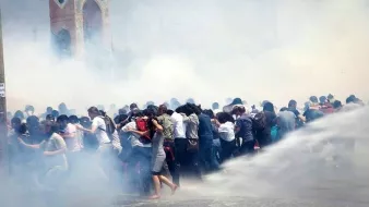 L'image montre une scène de manifestation où un grand groupe de personnes semble être en mouvement, probablement en train d'échapper à des gaz lacrymogènes ou à une intervention des forces de l'ordre. Le brouillard blanc, causé par la fumée ou les sprays, rend l'atmosphère tendue et chaotique. En arrière-plan, une structure architecturale est visible, mais elle est à peine discernable à cause de la fumée. Les manifestants semblent rassemblés et stressés, certains essayant de se protéger ou de se diriger vers une zone plus sûre.