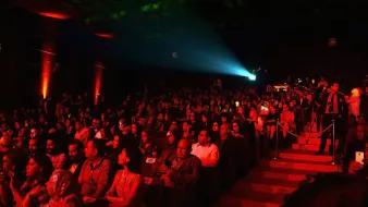 L'image montre un auditorium rempli de spectateurs. La salle est faiblement éclairée avec des lumières rouges qui créent une ambiance chaleureuse et dynamique. Au centre, un projecteur émet un faisceau lumineux, ajoutant à l'atmosphère du moment. Les spectateurs, composés d'hommes et de femmes, semblent attentifs et engagés, certains tenant des téléphones. L'escaliers sur le côté de la scène permet l'accès à d'autres niveaux. L'ensemble de la scène évoque un événement artistique ou culturel.