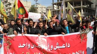 L'image montre un groupe de manifestants tenant des pancartes et des drapeaux, en train de défiler dans une rue. Ils portent des expressions déterminées et semblent militer pour une cause. Leurs vêtements et les drapeaux indiquent une provenance régionale. Une grande banderole rouge est affichée, avec des inscriptions en arabe. Les manifestants font le signe de la victoire avec leurs doigts, ce qui suggère un esprit de solidarité et de lutte. L'ambiance générale semble dynamique et engagée.