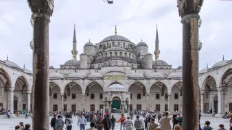 L'image montre une vue grandiose de la Mosquée bleue à Istanbul, encadrée par des arches majestueuses. Au premier plan, on peut voir un espace vaste et ouvert avec de nombreuses personnes qui se déplacent, créant une ambiance vivante. La mosquée elle-même se dresse en arrière-plan avec ses dômes et ses minarets élancés, sous un ciel nuageux qui ajoute une atmosphère particulière à la scène. Les détails architecturaux des colonnes et des arches sont également visibles, soulignant la beauté de cette œuvre historique.