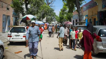 L'immagine mostra una strada affollata, probabilmente in un'area urbana. Ci sono diversi gruppi di persone che camminano, alcune sotto ombrelloni. Un uomo anziano con una canna si muove lentamente, mentre altri sembrano conversare e fare acquisti. Sullo sfondo, si possono vedere alcune automobili parcheggiate e alberi che offrono ombra. L'atmosfera sembra vivace e comunitaria, con il sole che illumina la scena.