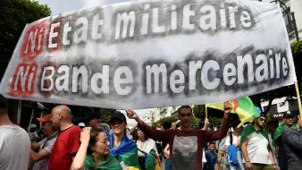 The image shows a large group of people participating in a protest or march. In the foreground, a man is holding a prominent banner that reads "Ni Etat Militaire Ni Bande Mercenaire," which translates to "Neither Military State Nor Mercenary Band." The crowd appears diverse, with individuals carrying flags and wearing various clothing styles, indicative of a collective demonstration. The background features urban scenery, suggesting the protest is taking place in a city.