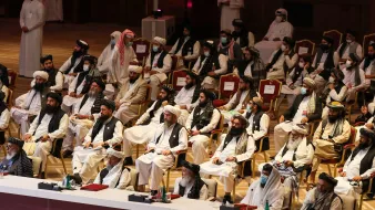 L'image montre un grand rassemblement de personnes assises dans une salle. Les participants sont principalement vêtus de vêtements traditionnels, tels que des turbans et des robes longues. Certains portent des masques. Les gens semblent attentifs et concentrés sur l'événement en cours. L'environnement est formel, avec des sièges disposés en rangées, et une scène à l'avant. L'atmosphère générale évoque une réunion officielle ou un congrès.