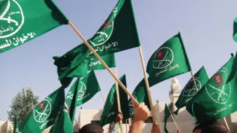 L'image montre un groupe de personnes brandissant des drapeaux verts. Les drapeaux portent probablement des symboles ou des inscriptions en arabe, ce qui suggère un contexte politique ou religieux. L'environnement semble être en extérieur, possiblement lors d'un rassemblement ou d'une manifestation.