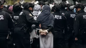 L'image montre un groupe de policiers en uniforme noir entourant une personne arrêtée. Cette personne est de dos, les mains attachées derrière. Les policiers portent des casques et des protections, et il y a une atmosphère tendue, suggérant une manifestation ou une intervention policière.