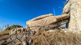 The image shows a large, partially fallen stone statue lying on its side against a rocky background, surrounded by sparse vegetation and a clear blue sky.