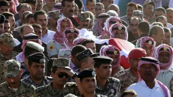 The image depicts a large group of people gathered in a public setting for a somber occasion. In the foreground, several individuals are wearing traditional attire, including headscarves and distinctive clothing, which suggests a cultural or regional significance. They appear to be carrying a casket draped in a flag, indicating a funeral or memorial service. The crowd encompasses military personnel dressed in uniforms, highlighting a sense of respect and ceremony. The atmosphere is likely one of mourning and solidarity as attendees pay their respects.