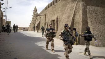 L'image montre une scène militaire dans une zone urbaine, probablement dans un pays du Sahel. Plusieurs soldats sont visibles, certains en uniforme camouflé et d'autres en tenue différente. Ils portent des armes et avancent dans une rue étroite bordée de bâtiments en terre battue, typiques de l'architecture sahélienne. On peut également apercevoir des civils en arrière-plan, ajoutant une dimension humaine à la scène. Le ciel est partiellement nuageux, ce qui suggère une ambiance chaude et aride.