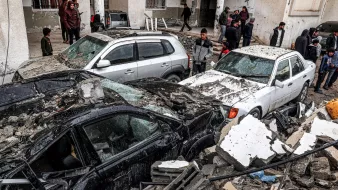 L'immagine mostra una scena di devastazione dopo un evento disastroso, probabilmente un terremoto. Si possono vedere diverse auto danneggiate, alcune sepolte sotto macerie e detriti. Il contesto è urbano, con edifici visibilmente danneggiati in fondo. Ci sono anche persone nelle vicinanze, che osservano la situazione, alcune sembrano preoccupate o increduli di fronte ai danni. La scena trasmette un forte senso di shock e di difficoltà.