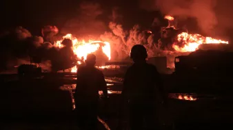 L'image montre deux silhouettes humaines marchant vers une scène d'incendie intense. De grands flammes et des nuages de fumée noire s'élèvent dans un environnement sombre, illuminé par la lumière des feux. Le sol semble humide, reflétant la lumière orangée des flammes.