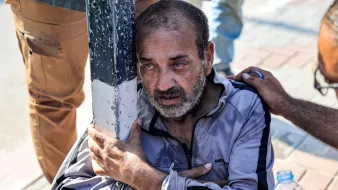 The image depicts a distressed elderly man sitting on the ground, clutching a pole for support. He appears visibly upset, with tears in his eyes and a look of despair on his face. His clothing is worn, and he seems to be in a tough situation. There are hands reaching out to him, suggesting someone is offering help or comfort. The background shows a pavement, indicating an outdoor setting. The overall atmosphere conveys a sense of vulnerability and struggle.