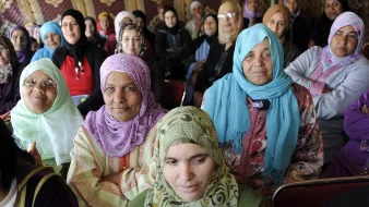 La imagen muestra a un grupo de mujeres, la mayoría de ellas vestidas con hijabs de diferentes colores y estilos. Ellas están sentadas en un ambiente que parece ser un evento o reunión, y sus expresiones varían entre la seriedad y la atención. Algunas mujeres llevan prendas tradicionales, y el fondo sugiere un entorno decorado con motivos culturales. La diversidad en los colores de los pañuelos y vestimenta resalta un sentido de unidad y diversidad entre ellas.