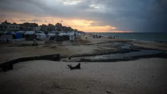 L'image montre une plage à l'heure du coucher de soleil, avec des nuages sombres dans le ciel. Au premier plan, on aperçoit du sable et une petite étendue d'eau stagnante. On peut également voir des tentes ou des abris, probablement des habitations temporaires, ainsi que quelques personnes se déplaçant sur la plage. L'atmosphère est à la fois paisible et mélancolique, avec des teintes orange et grises dans le ciel.