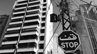 L'image montre une scène urbaine monochrome. À gauche, on aperçoit un bâtiment moderne avec plusieurs étages et balcons. À droite, un poteau électrique est visible, avec des fils enchevêtrés qui s'étendent dans le ciel. Il y a un panneau de signalisation en forme de hexagone avec le mot "STOP" écrit en arabe, ajouté à l'ambiance citadine. Le ciel est clair, ce qui donne une impression de luminosité à l'ensemble de la scène.