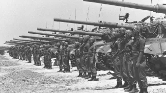 L'image montre un groupe de soldats alignés de manière formelle, portant des uniformes militaires et des casques. Ils sont positionnés devant une rangée de chars d'assaut, qui sont également alignés. Le paysage semble désertique, sous un ciel clair. L'atmosphère évoque une préparation militaire, peut-être pour une parade ou une démonstration de force. Les chars sont équipés de canons pointés vers l'avant, ce qui ajoute à l'impression de puissance et de militarisation de la scène.