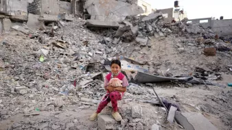 L'image montre une jeune fille assise sur des décombres, entourée de ruines. Elle porte des vêtements colorés et tient un jouet, semblant à la fois vulnérable et résiliente au milieu du chaos. La scène évoque des sentiments de tristesse et de perte, en raison de la destruction visible sur son environnement.