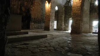 L'image montre un intérieur de cave ou de grotte, avec des murs en pierre apparente. On peut voir des arches qui soutiennent le plafond, et la lumière filtre par des fenêtres grillagées, créant une ambiance sombre mais fascinante. Le sol est en pierres irrégulières, et il semble y avoir un espace dégagé au centre. L'environnement dégage une atmosphère ancienne, presque mystique.