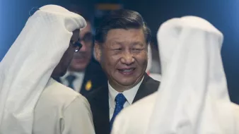 L'image montre un homme souriant, probablement un leader, en conversation avec deux autres personnes portant des turbans blancs. L'ambiance semble formelle et des éléments de l'environnement suggèrent un contexte officiel.