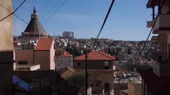 L'image montre un paysage urbain avec des bâtiments aux toits en tuiles rouges, typiques de certaines architectures méditerranéennes. On peut observer un dôme sur l'un des bâtiments, sans doute une église ou un édifice religieux. En arrière-plan, la ville s'étend sur plusieurs niveaux, avec de nombreuses constructions. Le ciel est dégagé et ensoleillé, créant une ambiance lumineuse. Des lignes électriques traversent l'image, ajoutant un élément urbain au paysage.