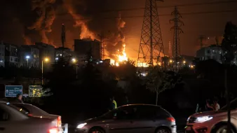Flammes illuminent la nuit, voitures passent près d'une scène de chaos urbain.