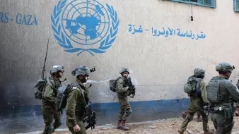 L'image montre des soldats en uniforme marchant près d'un bâtiment, probablement associé à l'ONU, avec un logo visible sur le mur. Leurs tenues militaires indiquent qu'ils sont en mission. L'environnement semble urbain, avec des structures typiques d'une zone habitée. L'atmosphère peut être perçue comme tendue en raison de la présence militaire.