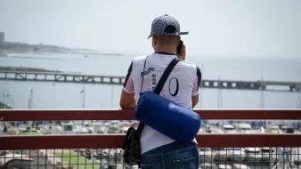 Un homme regarde la mer depuis un pont, avec un port et des bateaux en arrière-plan.
