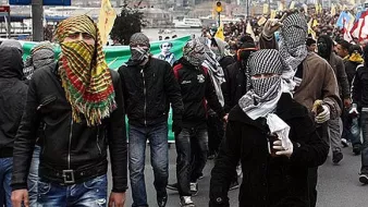 L'image montre une manifestation de foule, où de nombreuses personnes marchent ensemble. Beaucoup d'entre elles portent des masques ou des foulards qui couvrent leur visage, ce qui donne un aspect uniformisé à ce groupe. On constate une ambiance de protestation, probablement liée à une revendication sociale ou politique. En arrière-plan, on aperçoit des drapeaux et des banderoles, indiquant des messages forts. Les participants semblent déterminés, et l'atmosphère semble chargée d'énergie et de tensions.