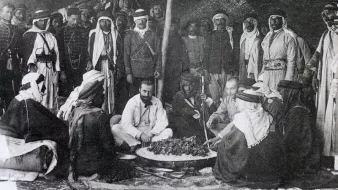 The image depicts a historical gathering of people, likely in a desert or rural setting. In the center, a group of men is seated around a large circular dish, which appears to contain food. They are dressed in traditional attire, with some wearing turbans and others in military or colonial-style uniforms. The background features a tent or canopy, suggesting a communal or ceremonial atmosphere. The mood seems convivial, as the participants engage with one another, possibly sharing a meal or discussing important matters. Overall, the scene reflects cultural practices and social interactions from the past.