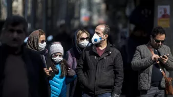 L'image montre un groupe de personnes marchant dans une rue. La plupart des individus portent des masques, ce qui indique qu'ils respectent des mesures de santé, probablement en raison d'une pandémie. Parmi eux, une jeune fille porte une veste bleue et des lunettes. Les adultes sont vêtus de manière variée, avec des manteaux et des écharpes. L'atmosphère semble être celle d'une ville, avec des bâtiments flous en arrière-plan.