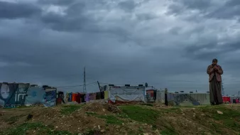 L'image montre une scène en extérieur sous un ciel nuageux. À droite, un homme se tient debout, apparemment pensif, avec une main près de son visage. En arrière-plan, on voit une rangée de tentes ou abris, probablement dans un camp, avec des murs en toile de différentes couleurs. Le sol est herbeux et légèrement surélevé. L'atmosphère est sombre en raison des nuages menaçants qui couvrent le ciel.