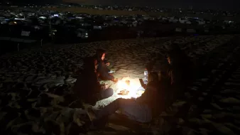 L'immagine mostra un gruppo di persone sedute intorno a un piccolo fuoco in una zona desertica durante la notte. Il terreno è sabbioso e la luce del fuoco crea un'atmosfera calda mentre il resto del panorama è buio. Sullo sfondo si possono vedere delle tende, probabilmente un accampamento, che sono illuminate da luci lontane. Le persone sembrano impegnate in una conversazione o in attività condivise, avvolte in abbigliamento caldo per proteggersi dal freddo notturno.