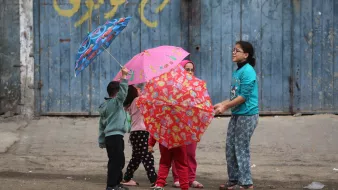 L'immagine mostra un gruppo di bambini che giocano insieme in strada, mentre tengono in mano degli ombrelli colorati. I bambini indossano abiti semplici e variopinti, e sembrano divertirsi sotto un muro blu con scritte. La scena trasmette gioia e convivialità.