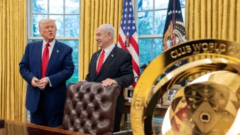 Deux hommes souriants devant un bureau, drapeaux en arrière-plan, avec un trophée doré au premier plan.
