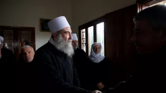 Group of men, some with white hats and beards, shaking hands and wearing dark clothing in a room with windows.
