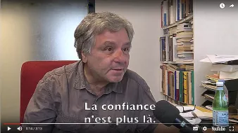 L'image montre un homme assis devant un bureau, dans un bureau plutôt ordonné avec des livres en arrière-plan. Il parle dans un microphone et semble discuter d'un sujet important. Sur l'écran, on peut voir le sous-titre qui indique : "La confiance n'est plus là." L'éclairage est neutre, et l'homme a une expression sérieuse.