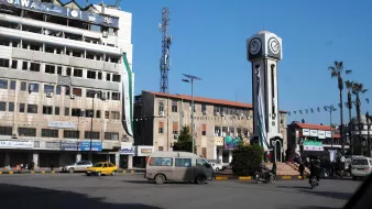 Una piazza con un grande orologio, edifici e alcuni veicoli in movimento.