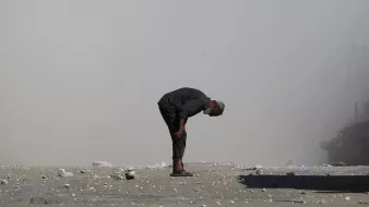 A man bends over on a dusty street, surrounded by debris and smoke.