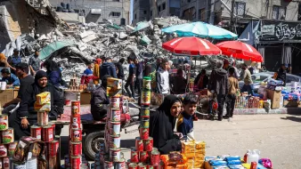 The image depicts a busy street scene in an area that appears to have experienced significant damage, as evidenced by the rubble and destroyed buildings in the background. In the foreground, a woman wearing a black hijab is sitting beside a young boy. They are surrounded by stacks of canned goods and other products that appear to be for sale, indicating a market or makeshift shop. People are seen walking through the street, some engaging in commerce amid the chaotic surroundings. Colorful umbrellas, likely providing shade, add contrast to the scene. Overall, it captures a moment of resilience and daily life amid challenging conditions.