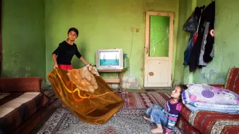 L'image montre une pièce modeste avec des murs verts. Au centre, deux enfants sont présents. L'un d'eux, un garçon, tient une grande couverture qu'il semble s'apprêter à lancer ou à étendre. L'autre enfant, une petite fille, est assise sur le sol, regardant le garçon avec curiosité. On peut voir un téléviseur allumé sur une petite table, et quelques vêtements accrochés au mur. Le décor est simple, avec un tapis et des canapés. L'ambiance générale semble familiale et décontractée.