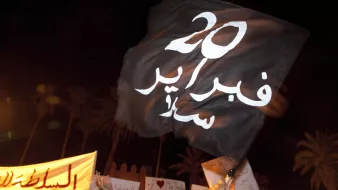 L'image montre une manifestation nocturne. À l'avant-plan, une personne tient un drapeau noir portant des inscriptions en blanc. Le drapeau semble être un symbole de protestation. En arrière-plan, on distingue des panneaux de revendications, également en arabe. L'atmosphère semble dynamique et engagée, illustrant un mouvement de contestation. La présence des palmiers suggère que l'événement se déroule dans un pays à climat chaud, probablement en Afrique du Nord.