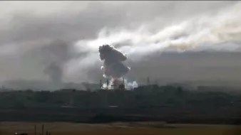 L'image montre une scène dramatique avec des nuages de fumée épais s'élevant dans le ciel, probablement à la suite d'une explosion ou d'un bombardement. On peut voir des silhouettes d'infrastructures industrielles ou urbaines partiellement obscurcies par la fumée. L'atmosphère est sombre et chargée, suggérant des conditions de conflit ou de destruction. La lumière semble faible, ajoutant à l'impression de chaos et de détresse.