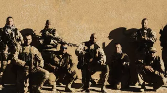 La imagen muestra a un grupo de soldados en un entorno desértico. Estos hombres están vestidos con trajes de combate y llevan equipamiento militar, incluyendo armas. Están organizados en una formación, posando para la foto. Algunos tienen gafas de sol y están en una posición que sugiere preparación y unidad. El fondo es simple, con una pared de color claro que resalta a los soldados.